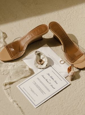Tan clear-strap heeled mules beside a wedding invitation, gold rings, and seashells on a sandy textured surface.