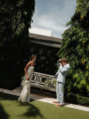 Groom in light blue suit photographs bride in white gown outdoors near lush green trees at elegant wedding venue.