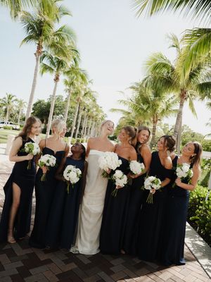 Bride in white gown with bridesmaids in navy dresses holding white bouquets, laughing outdoors among tropical palm trees.