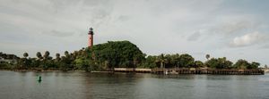 Panoramic view of Jupiter Inlet Lighthouse surrounded by lush green trees reflecting on calm blue waters in Florida.
