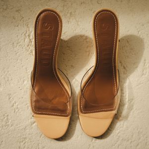 STAUD beige leather slide sandals with clear strap and block heel, shown from above on a sandy textured surface.