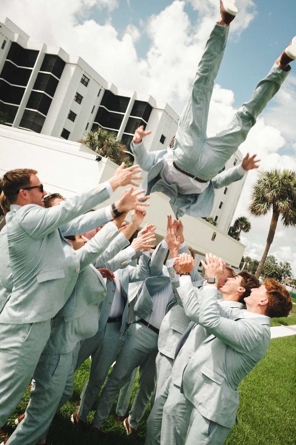 Groomsmen in light blue suits toss the groom into the air outdoors on a sunny wedding day.