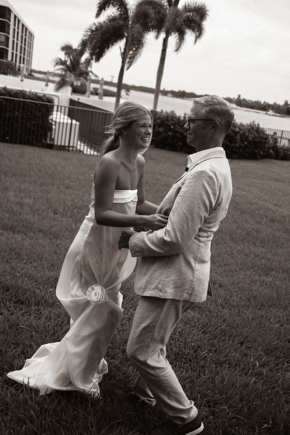 Black and white photo of a smiling bride in white gown and groom in light suit dancing on grass with palm trees.