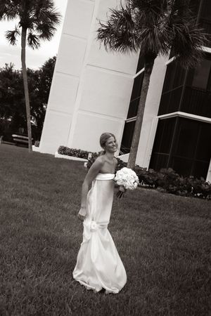 Bride in white mermaid gown holding bouquet smiling outdoors near palm trees and white building in black and white photo.