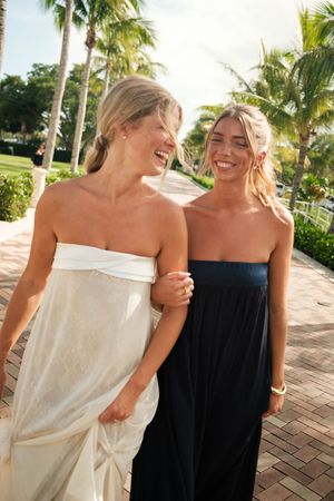 A laughing bride in white gown walks arm-in-arm with a friend in a black dress along a tropical palm-lined pathway.