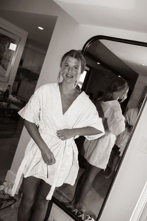 Woman in white robe smiling near a mirror in a room, captured in black and white photography.