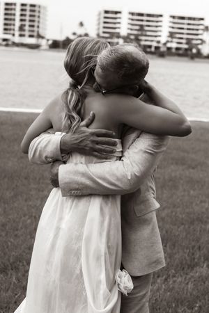 Black and white photo of a bride and groom sharing a tender embrace outdoors near a waterfront on their wedding day.