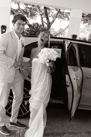 Black and white photo of a laughing bride holding bouquet and groom in suit arriving at wedding venue by car.