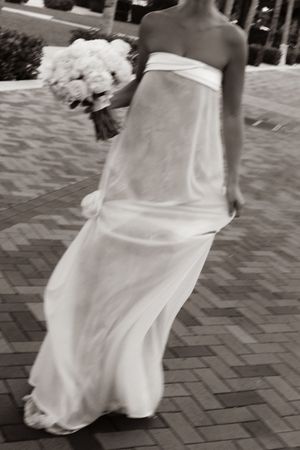 Bride in strapless white gown holding bouquet walking on brick path, black and white wedding photo.