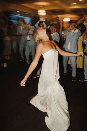 Bride in white gown dancing joyfully at wedding reception surrounded by cheering guests.