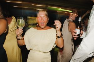 Woman in white dress dancing at party, holding wine glass and cigarette, wearing gold bracelets.
