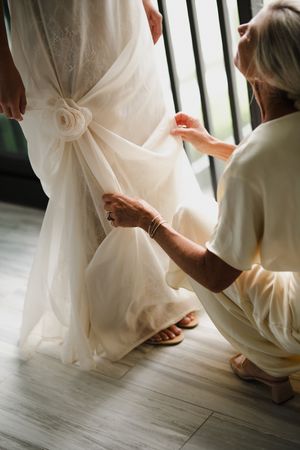 Mother kneeling to adjust bride's flowing white wedding dress train before ceremony.