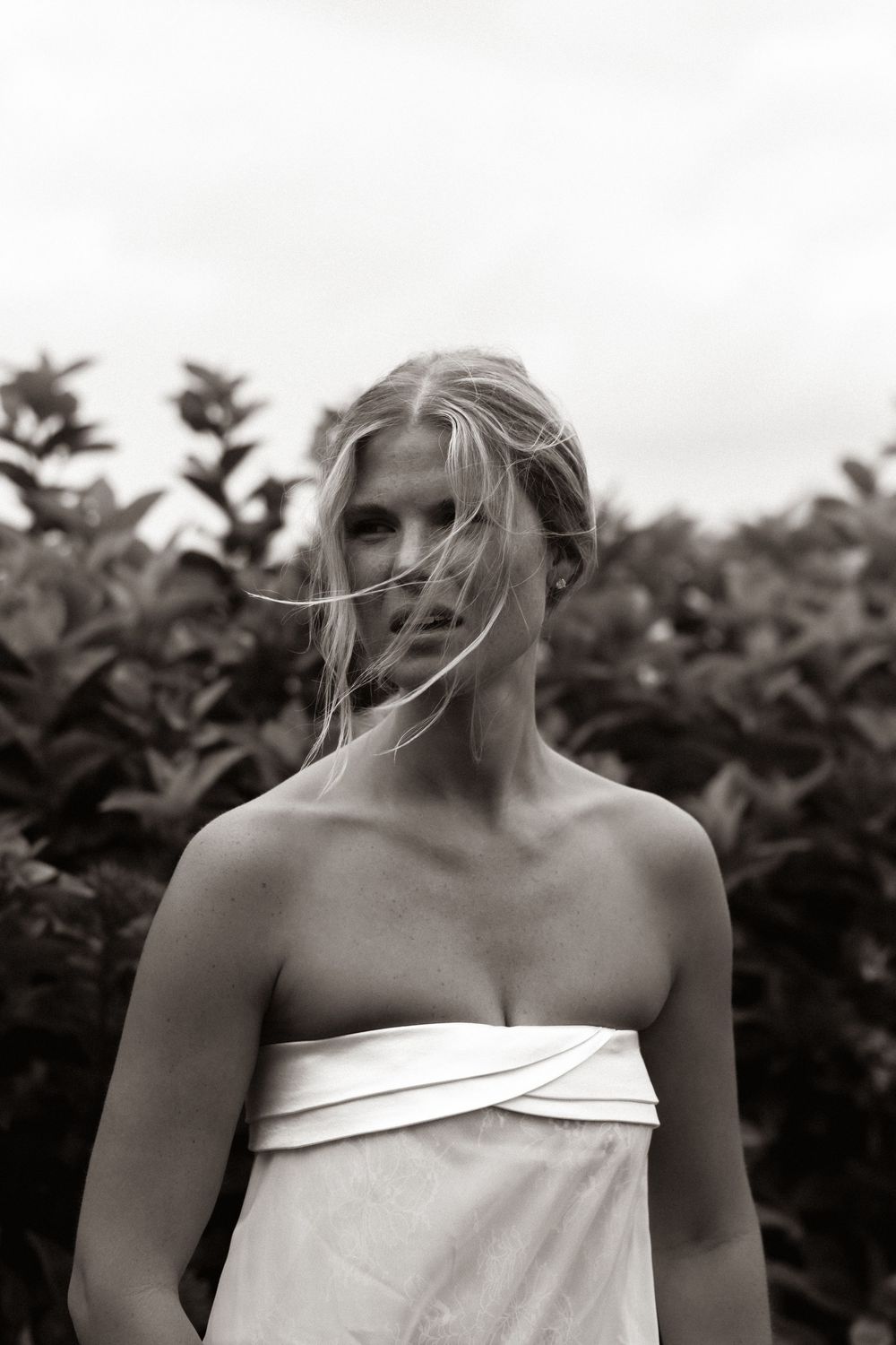 Black and white portrait of a blonde woman in a white strapless dress standing outdoors near lush green bushes.