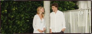 A couple in white outfits smiling at each other outdoors near green hedges and a white fence.
