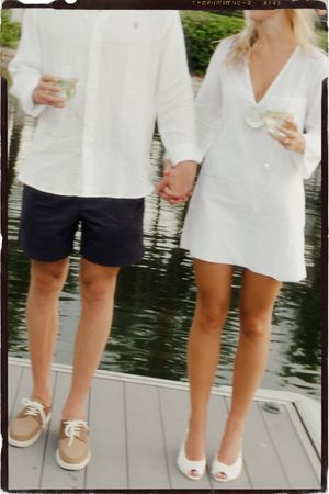 Couple holding hands on a dock, wearing white tops, man in navy shorts with tan loafers, woman in white mini dress with heels.