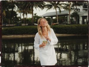 Smiling blonde woman in white shirt holding a drink near a tropical waterway with palm trees.