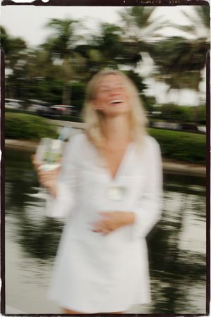 Blurred motion photo of a laughing blonde woman in a white dress holding a drink near a tropical waterway.
