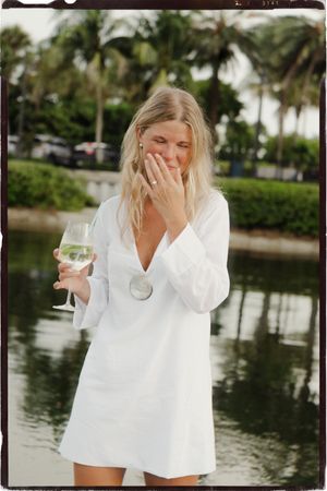 Blonde woman in white mini dress laughing, holding wine glass near tropical waterway with palm trees.