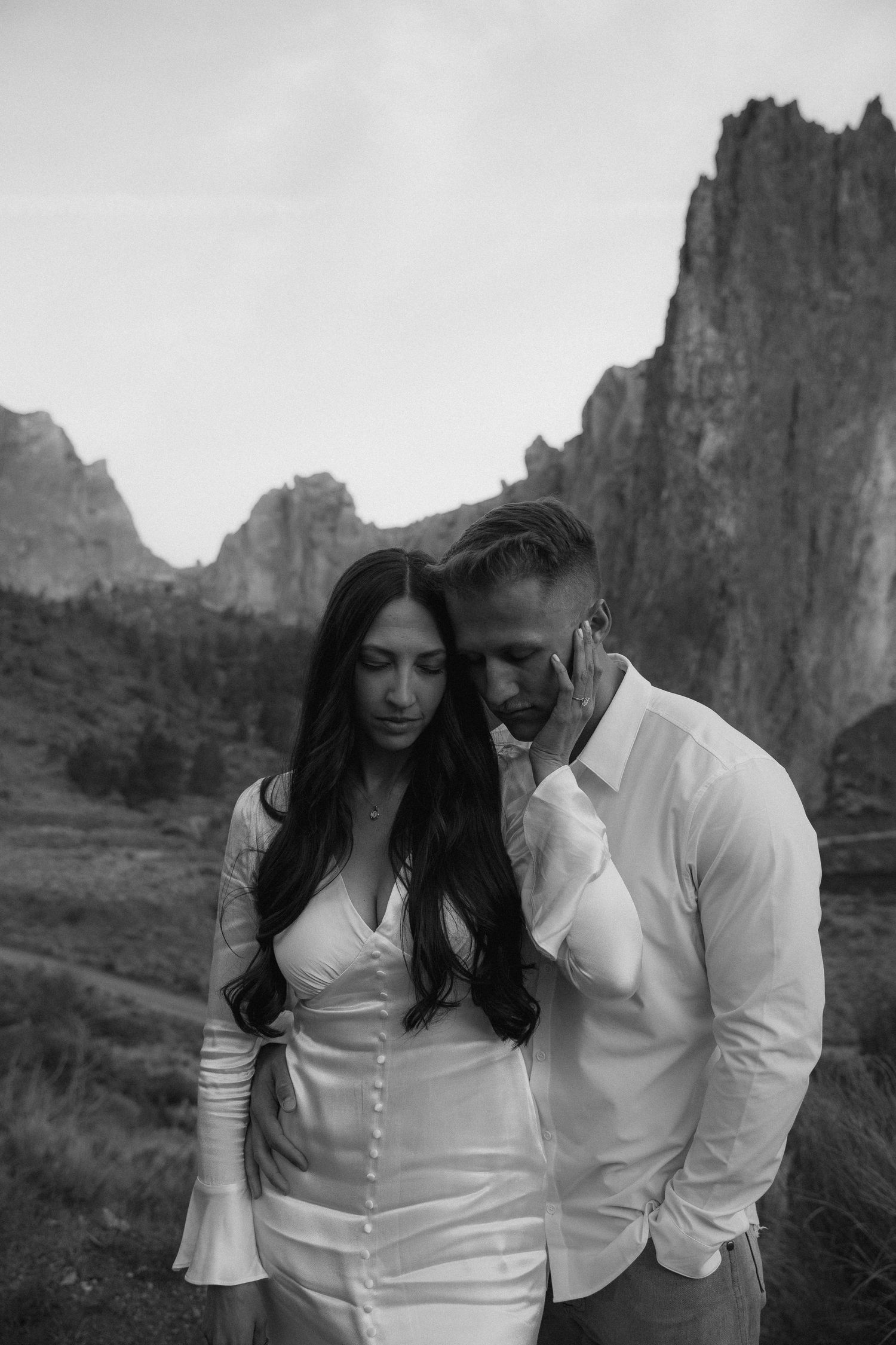 A couple in elegant white attire stands together against a dramatic backdrop of Smith Rock State Park in black and white.