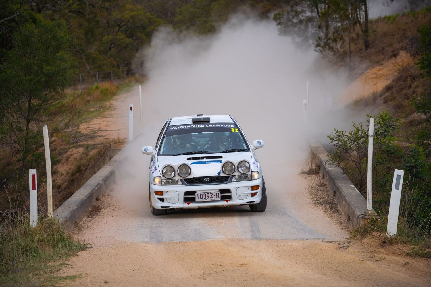 Rally car kicking up dust while crossing a small bridge on a dirt road.