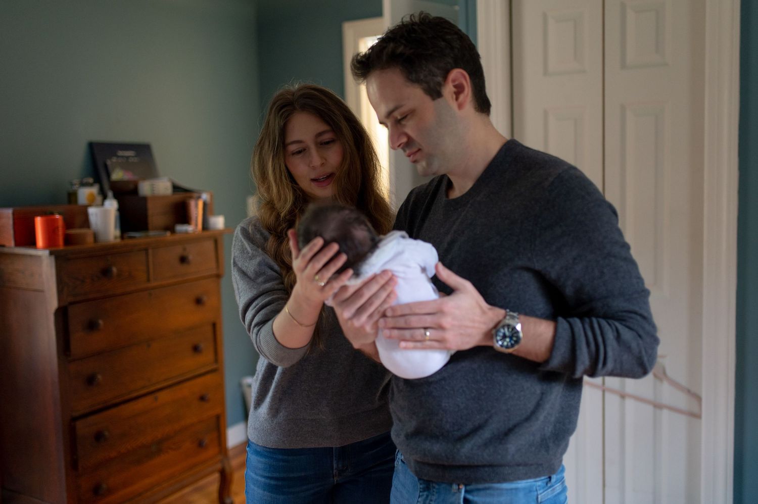 New parents holding their baby in a room with wooden furniture.