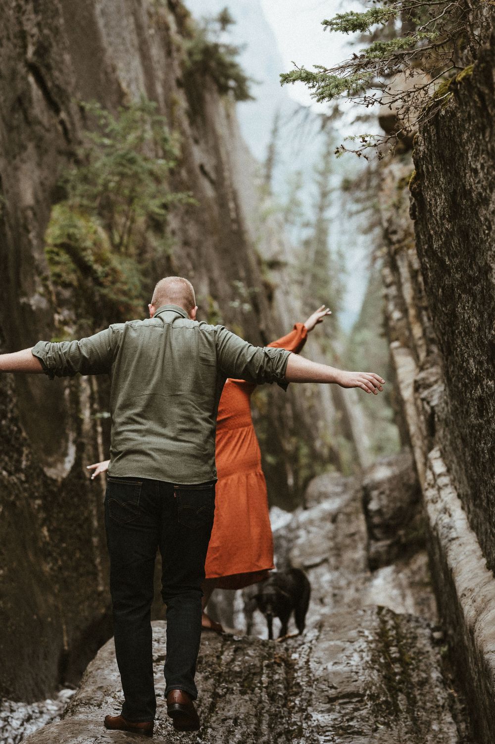 Two people in green and orange outfits walking along a rocky mountain path with arms spread wide.