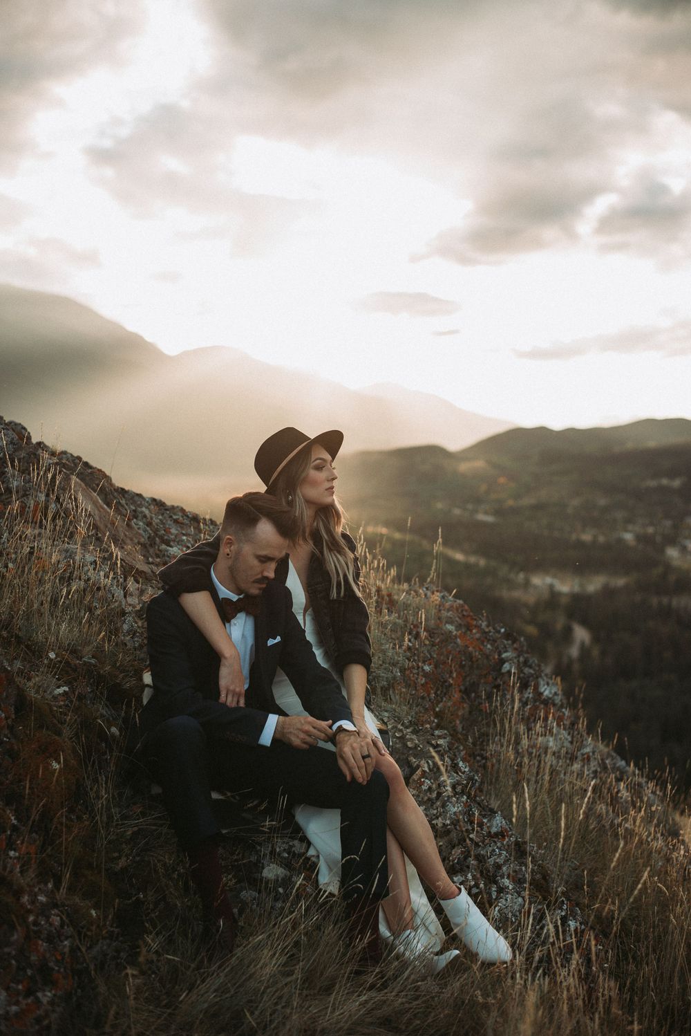 A couple relaxes on a mountainside during sunset, with dramatic valley views below.