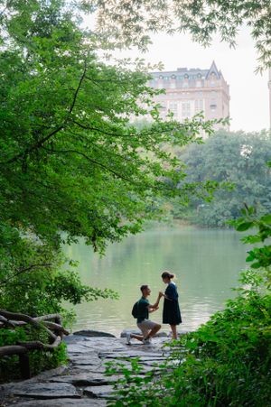 A romantic lakeside proposal scene in Central Park with a historic building visible in the background.