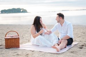 A couple enjoys a romantic beach picnic on a white blanket with wicker basket at sunset.
