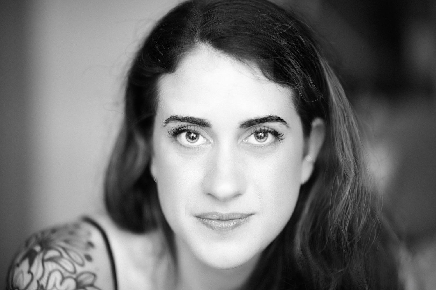Black and white portrait photograph showing dark wavy hair, gentle smile and visible shoulder tattoo against light background.
