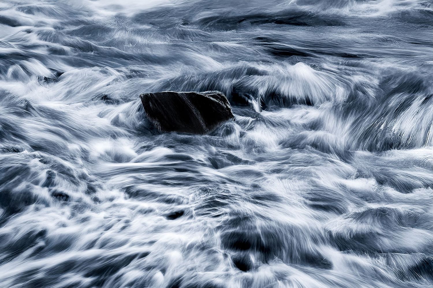 A dark rock surrounded by flowing water creates dramatic motion blur patterns in blue and white tones.