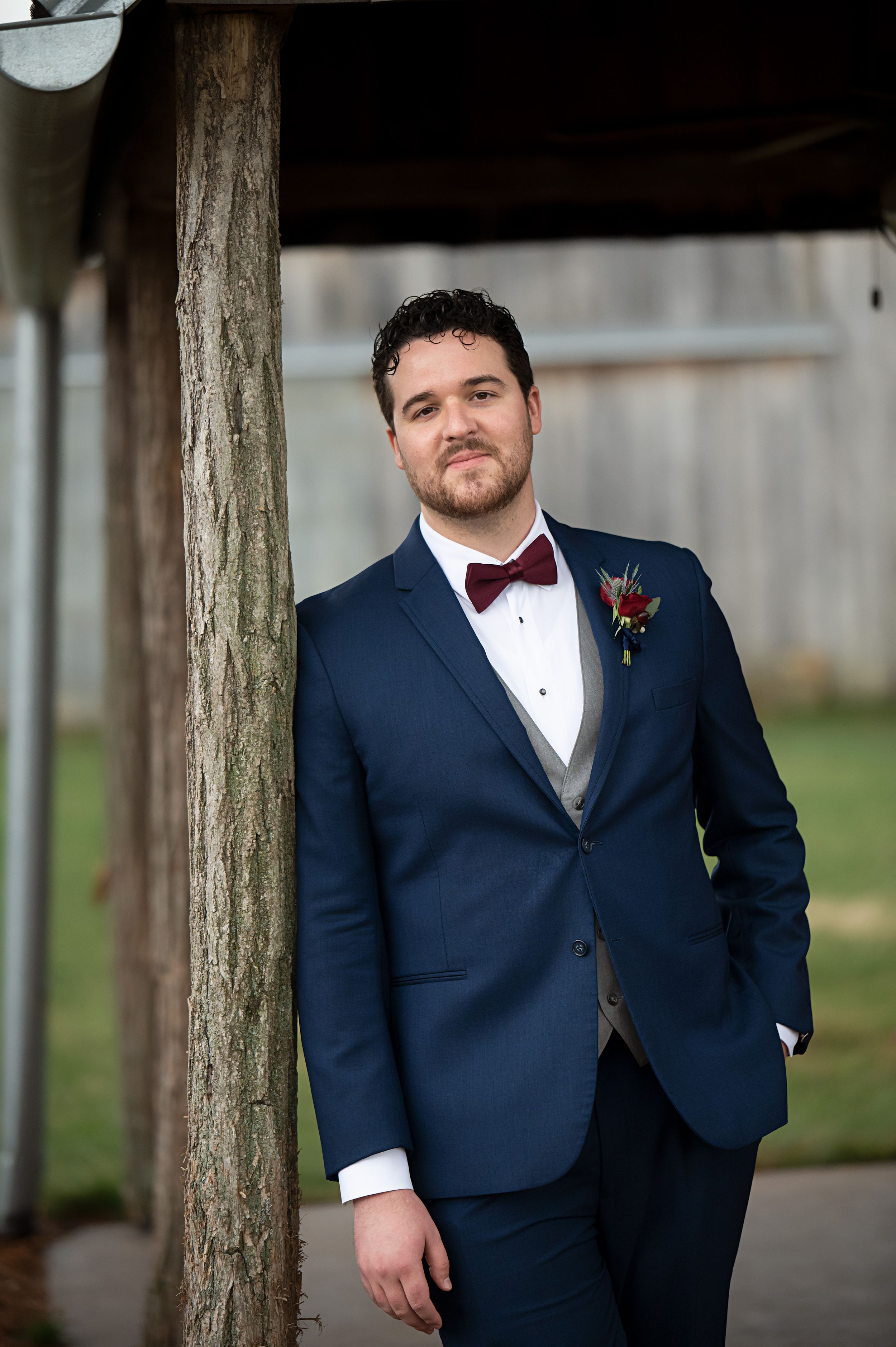 Groom in navy blue tuxedo poses between wooden posts for formal wedding portraits.