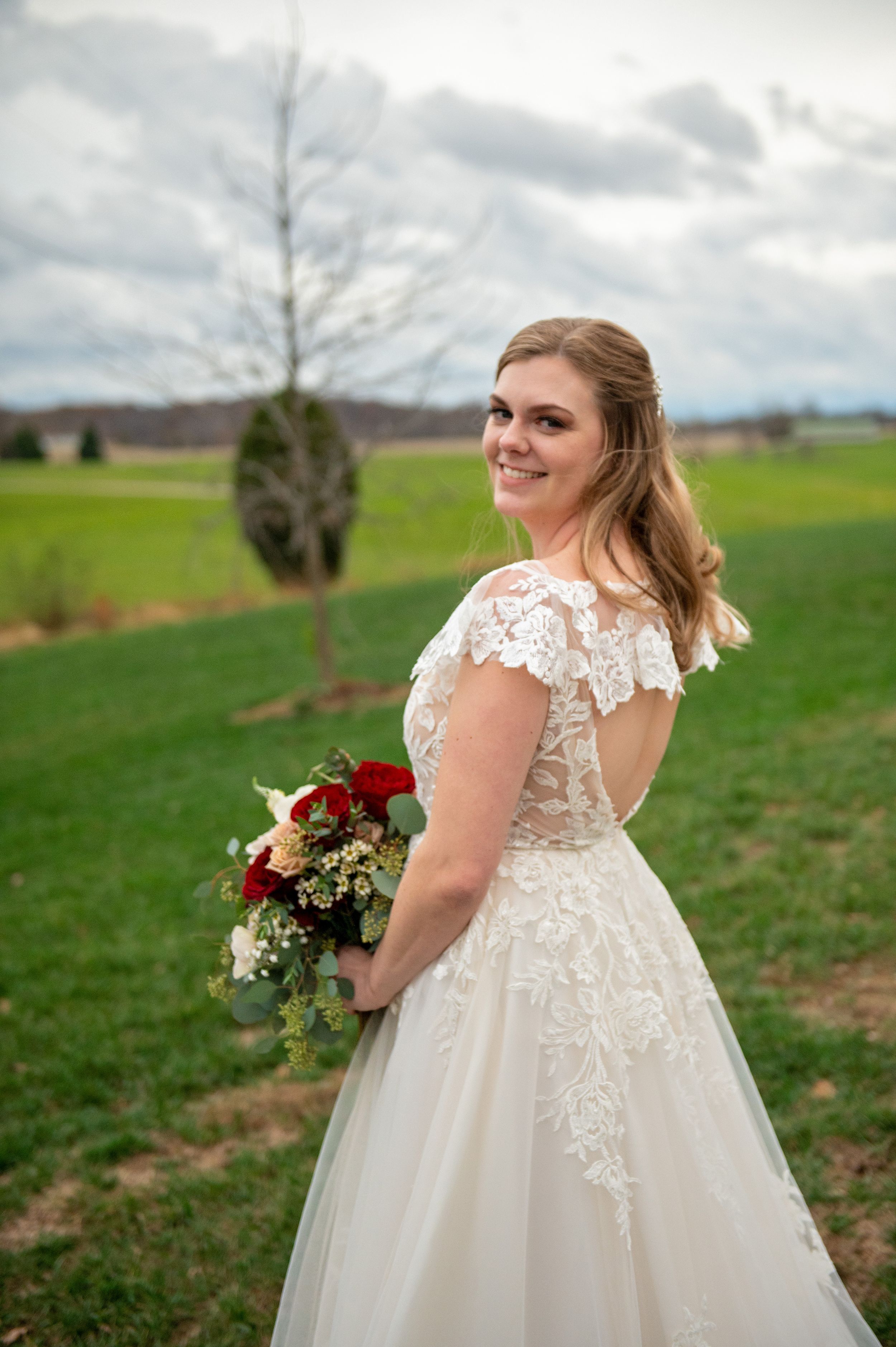 Bride in lace cap sleeve gown poses in countryside setting holding red bouquet.
