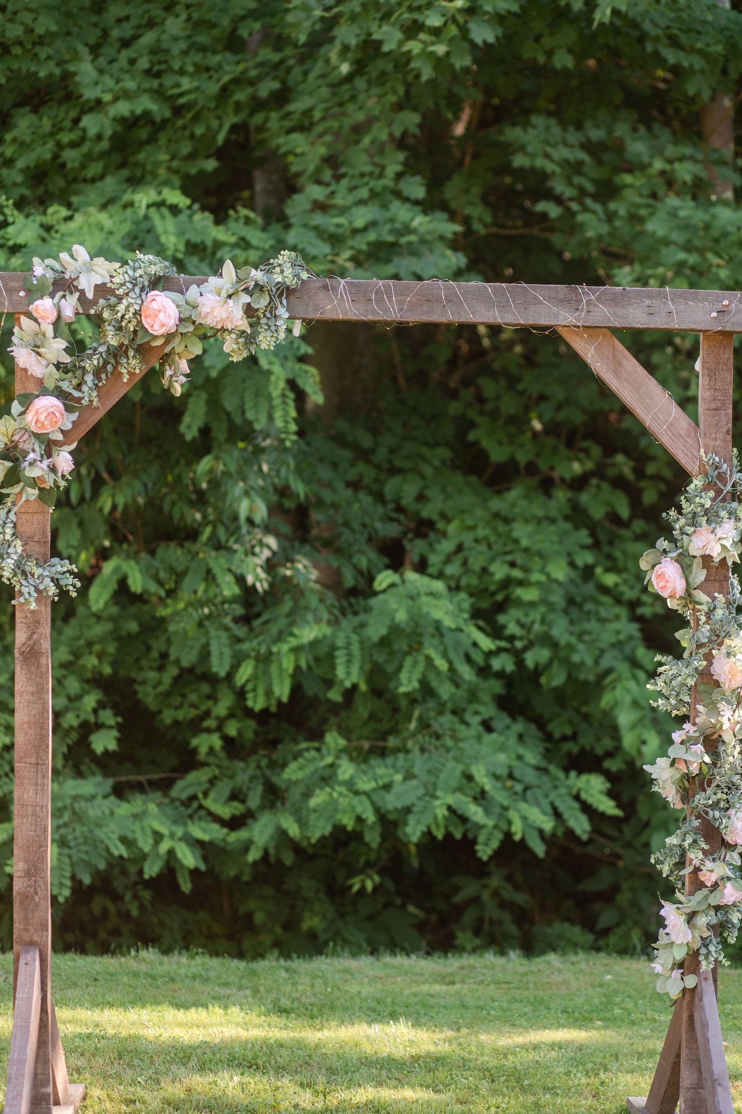 Wooden wedding arch decorated with pink and white flowers and greenery in an outdoor garden setting.