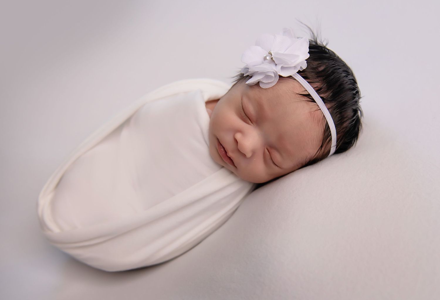 Newborn baby sleeping peacefully wrapped in white swaddle with matching headband.