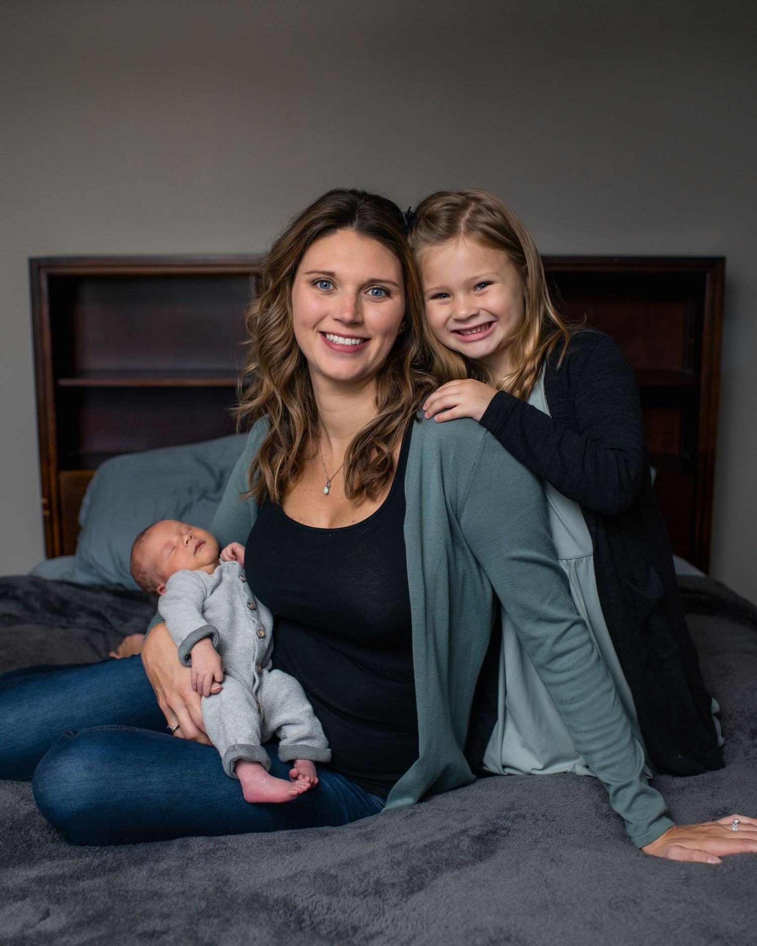 Family portrait on gray bed featuring a newborn baby and two others in casual attire.