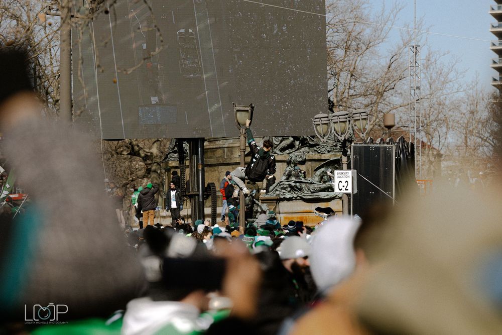 Outdoor crowd of people looking in one direction while standing beneath bare winter trees along an urban street.