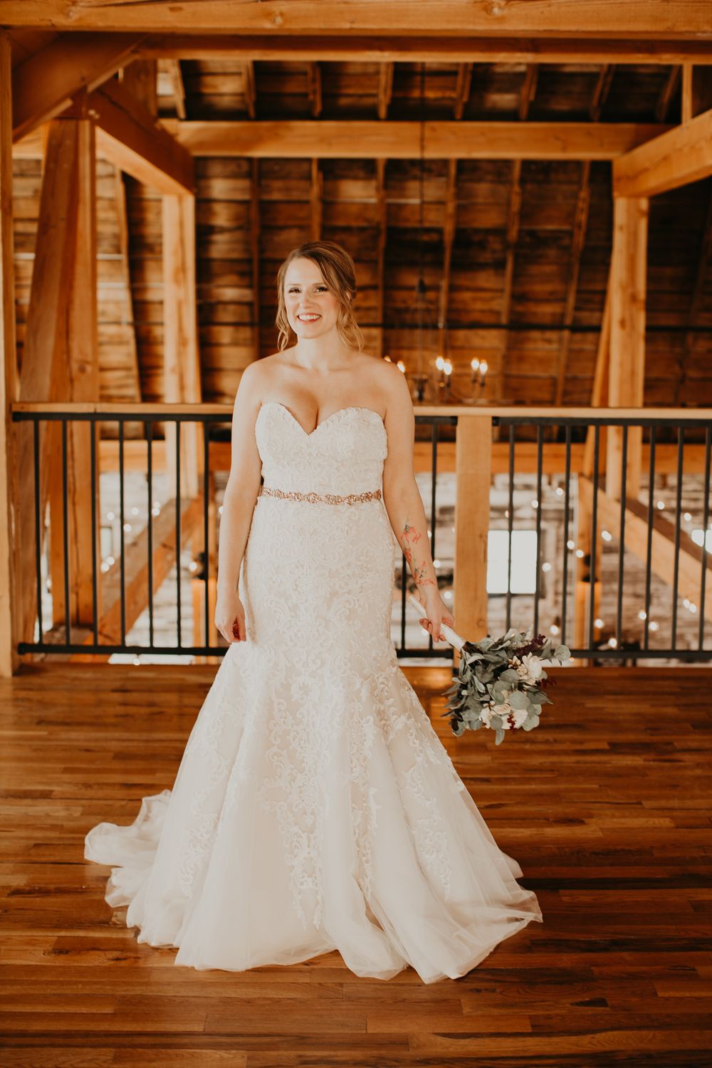 Bride in strapless lace mermaid gown holds bouquet inside rustic wooden barn wedding venue.