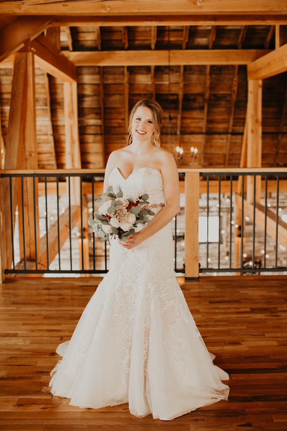 Smiling bride in lace mermaid wedding dress holds floral bouquet in elegant rustic barn venue.