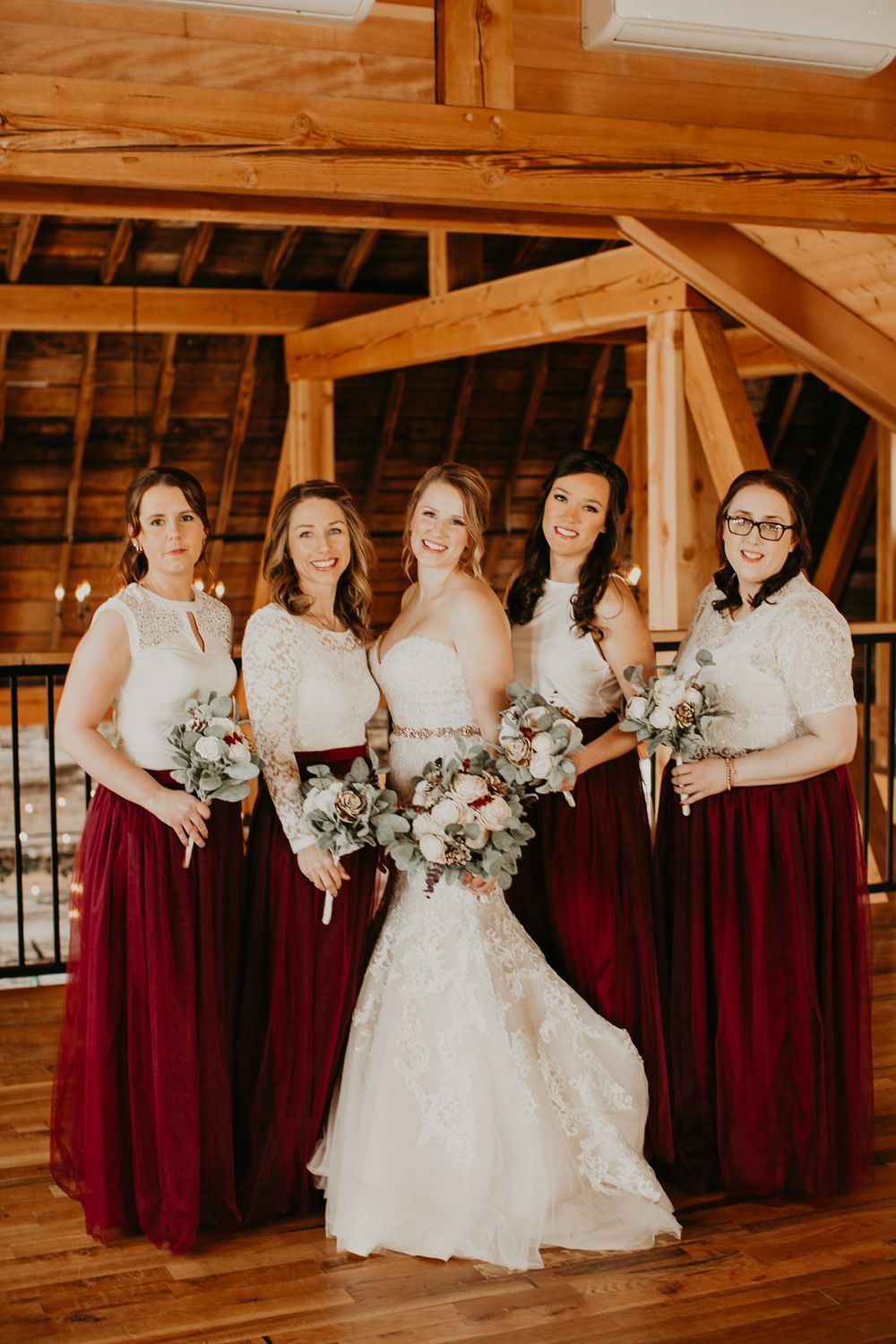 Bride in lace mermaid gown poses with four bridesmaids in white tops and burgundy skirts in barn.