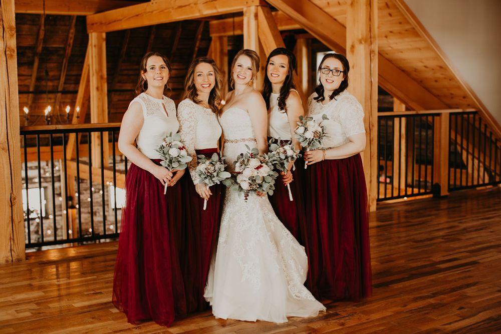 Bride in mermaid gown smiles with four bridesmaids in white lace tops and burgundy skirts in barn.