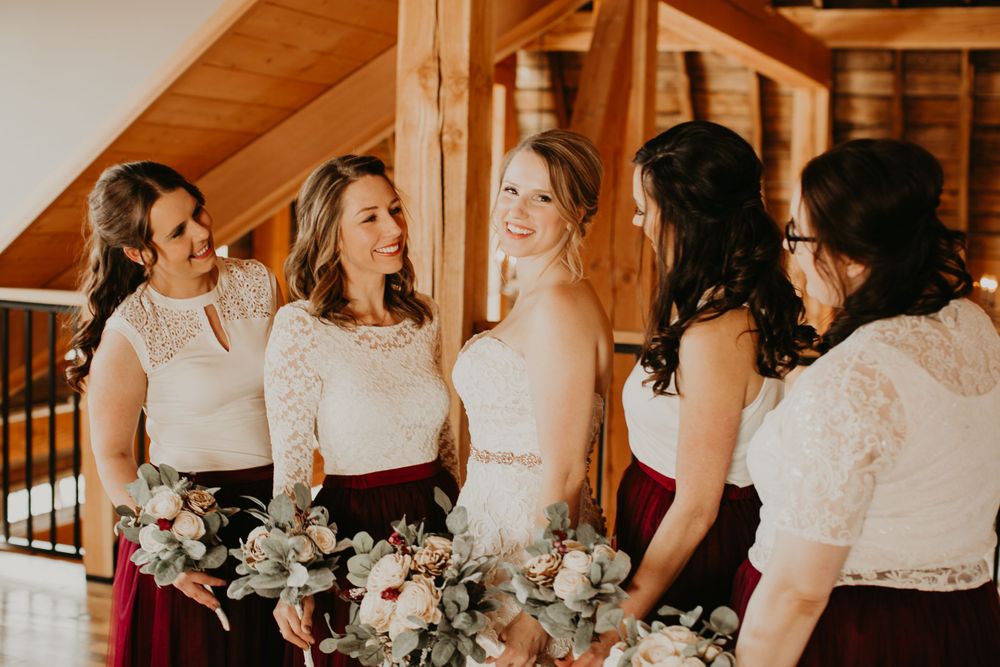 Bride and bridesmaids in burgundy and white laugh together holding bouquets in rustic barn venue.