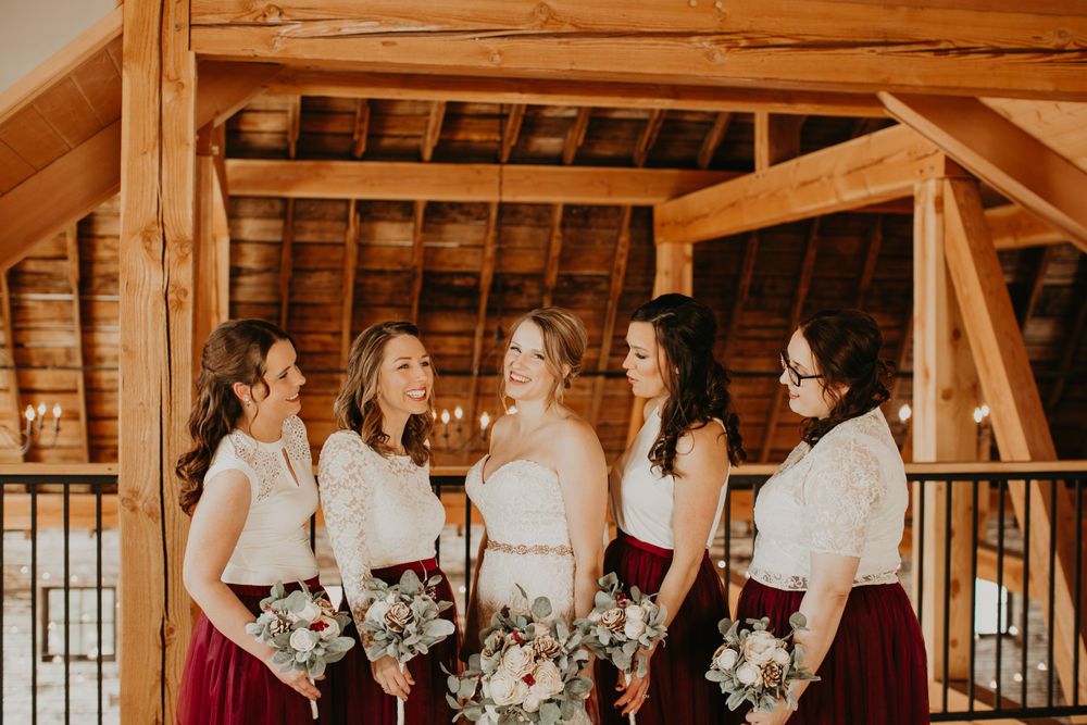Bride and bridesmaids in burgundy and white laugh together in a warmly lit rustic barn setting.