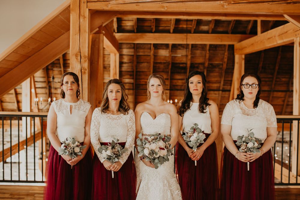 Bride and four bridesmaids in matching burgundy skirts pose seriously in rustic wooden barn venue.