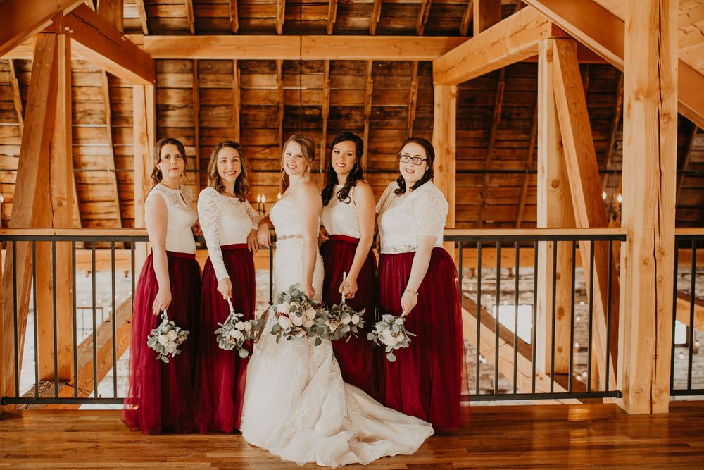 Bride in white mermaid gown poses with bridesmaids in burgundy skirts on rustic barn balcony.