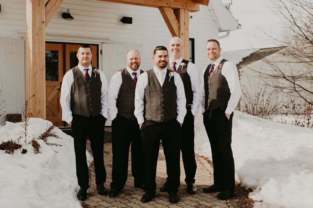 Five groomsmen in matching gray vests and dark pants pose outdoors in winter snow at a wedding venue.
