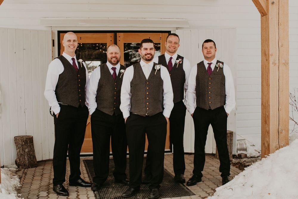 Groomsmen in gray vests and white shirts stand together in front of wooden barn doors in winter.