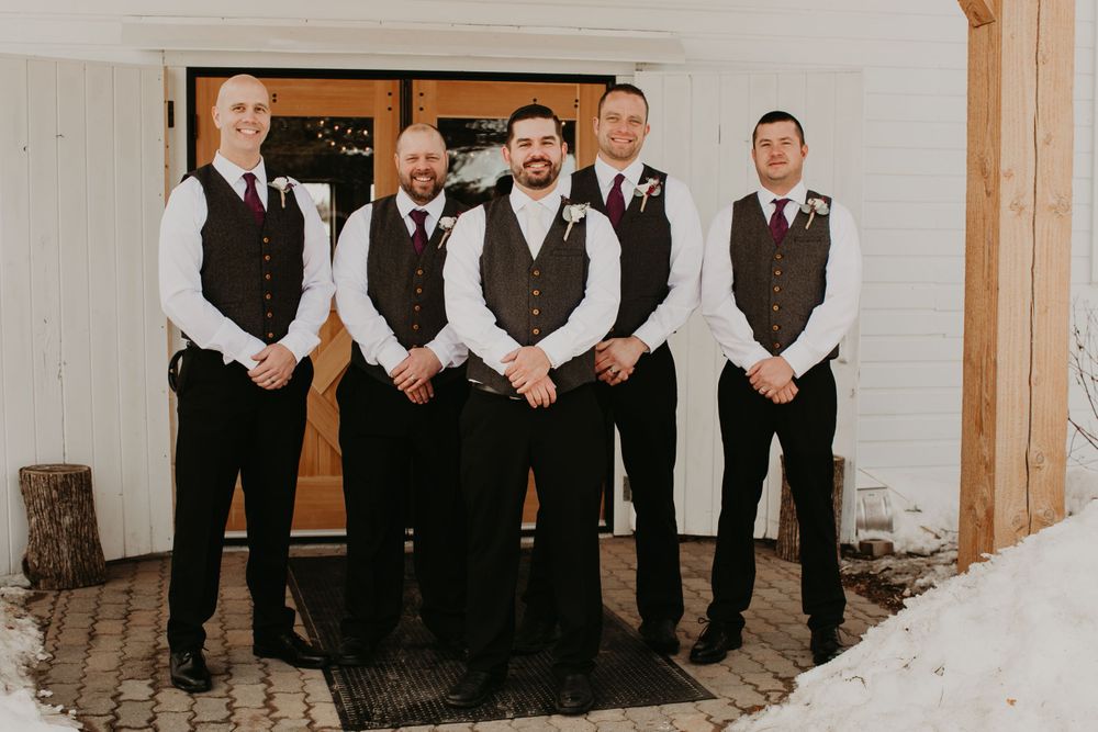 Five groomsmen wearing gray vests and burgundy boutonnieres pose at a rustic winter wedding venue.