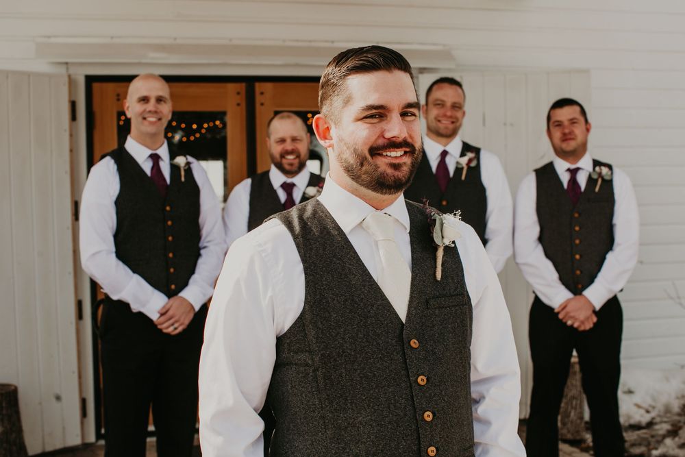 Groom in gray herringbone vest with boutonniere stands before his groomsmen at a winter wedding.