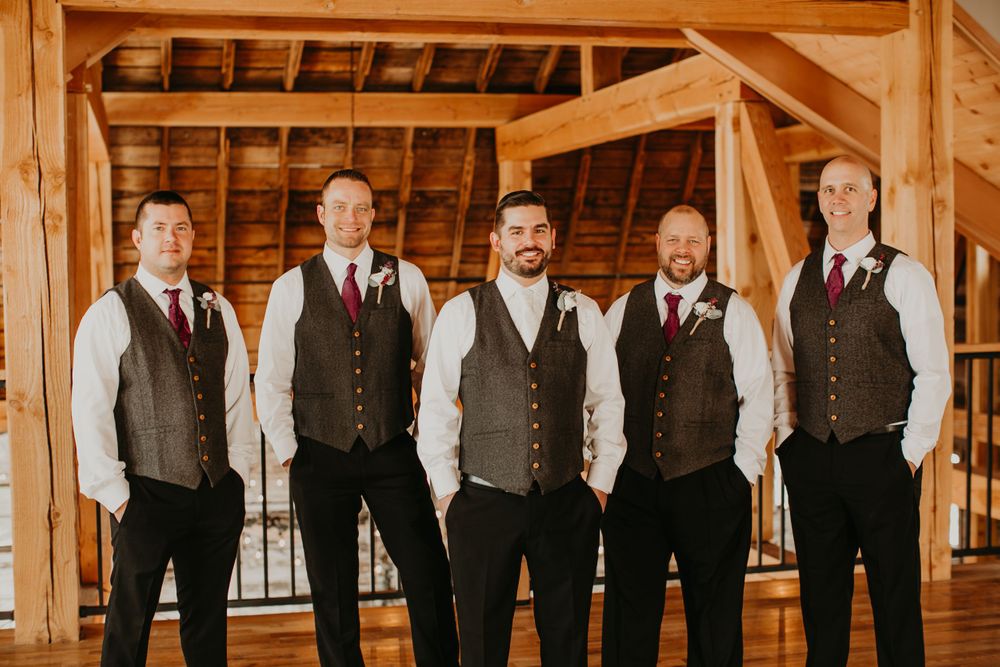 Five groomsmen in gray vests and dark pants pose inside a rustic wooden barn wedding venue.